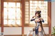 A woman in traditional attire stands near a window with wooden frames and a plant beside her.